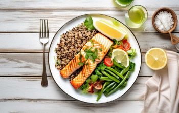 Top-down view of a colorful pescatarian meal with grilled salmon, quinoa salad, green beans, and lemon on a white shabby wooden table.